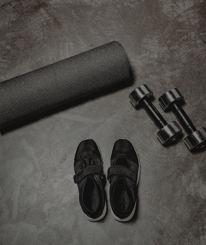 Black training shoes beside grey dumbbells and a black fitness mat on a dark grey gym floor.